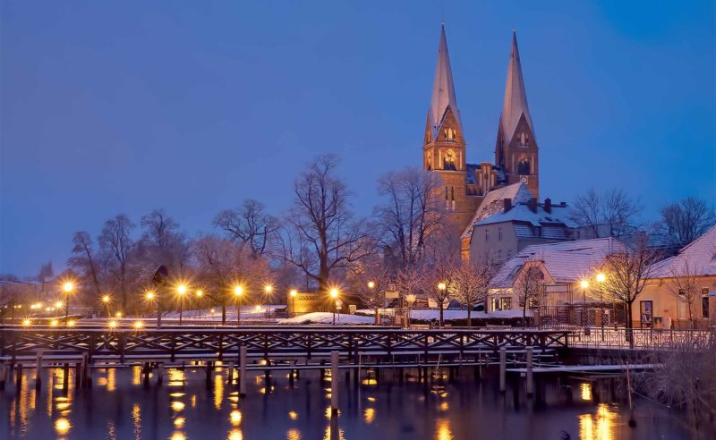 Region Neuruppin mit schneebedeckten Dächern und spiegelnden Lichtern im See bei Dämmerung