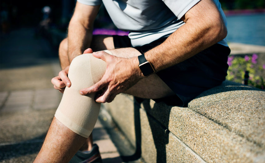 Mann sitzt auf Mauer und begutachtet sein Knie mit Bandage