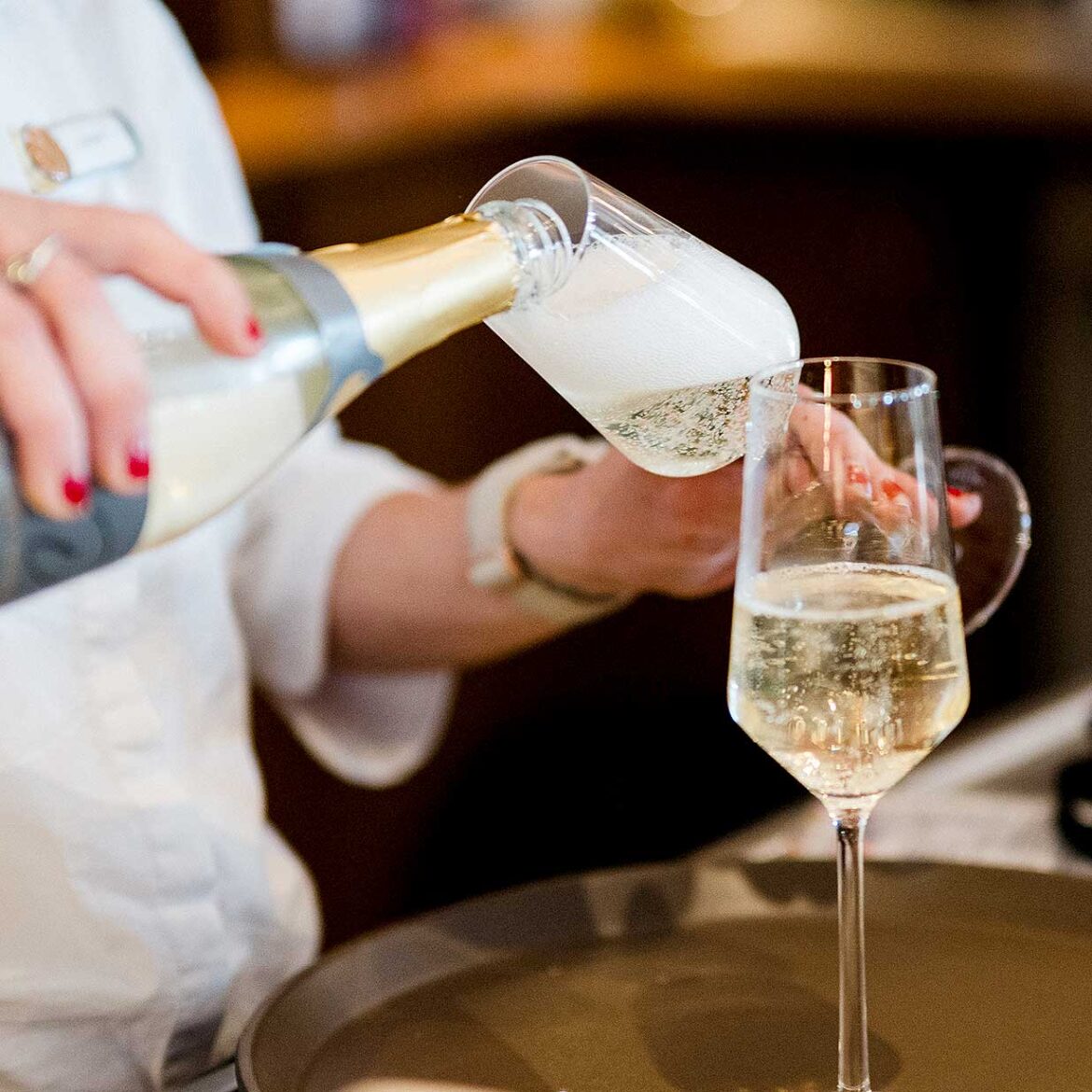 Nahaufnahme: Servicekraft schenkt perlenden Sekt aus einer Flasche in ein Sektglas auf einem Tablett ein