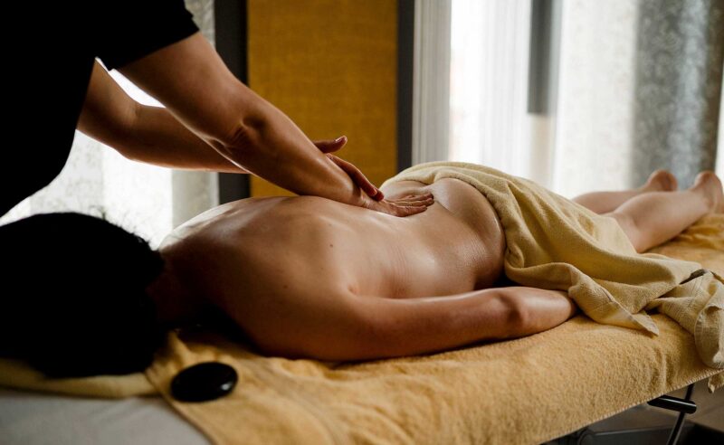 Therapeutin massiert den unteren Rücken einer Frau auf einer Massageliege, ein schwarzer Hot-Stone liegt im Vordergrund