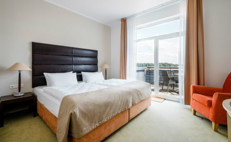 Blick auf ein großes Doppelbett, einen Sessel in orange sowie eine Tür zum Balkon.