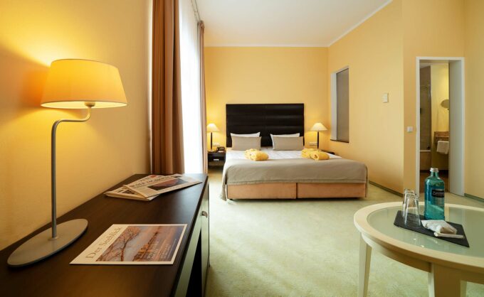 Helles Hotelzimmer mit Doppelbett, Tisch, Kommode und Blick ins Badezimmer.
