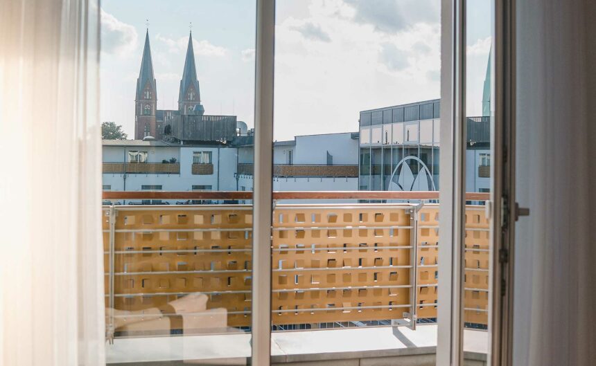 Offene Balkontür mit Blick über den Balkon zu einer Kirche mit zwei spitzen Türmen.