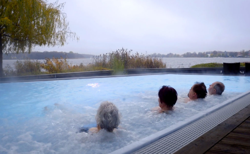 Vier Hotelgäste genießen die Aussicht auf den See im beheizten Außenpool der der Fontane Therme in Brandenburg.