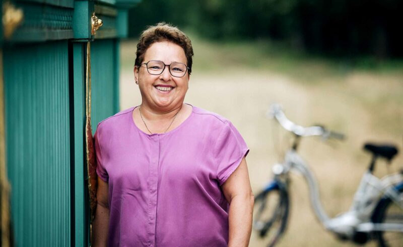 Nahaufnahme einer lächelnden Frau mit pinkem Oberteil, im Hintergrund steht ein Fahrrad.