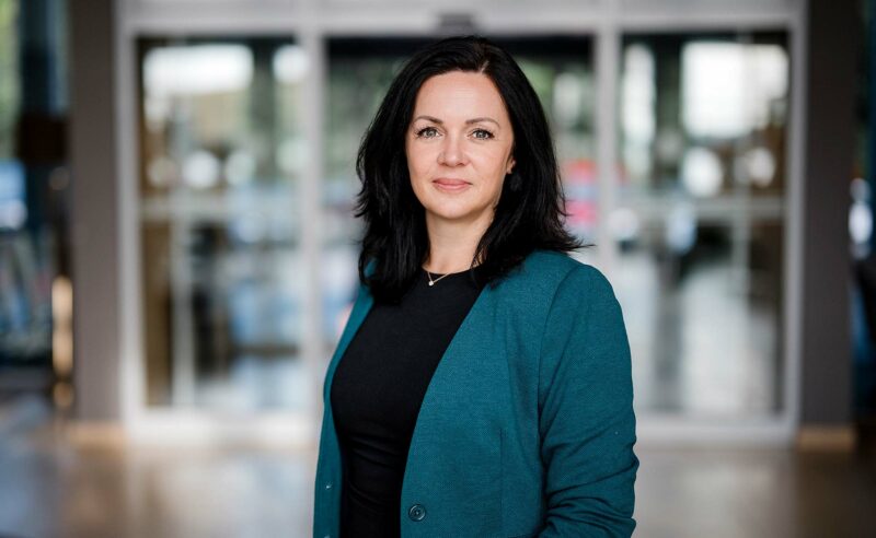 Portrait einer lächelnden Frau mit schwarzen Haaren, Oberteil sowie türkisem Cardigan.